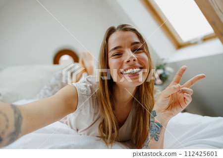 A woman on a bed, flashing a smile and peace sign while taking a selfie 112425601