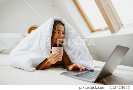 A woman is comfortably laying in bed under her blanket, with a cup of coffee and her laptop 112425635