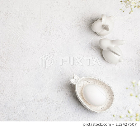 Minimal monochrome white Easter card with egg, ceramic bunnies, Gypsophila flowers on top. Top view 112427507