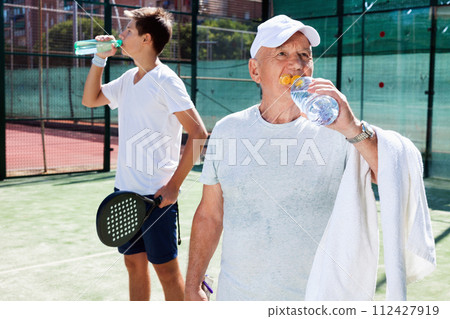 Padel players of different generations drink water on padel court Padel players of different generations drink water on padel court 112427919