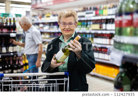 Mature woman with glasses chooses bottle of wine in alcohol section of supermarket Mature woman with glasses chooses bottle of wine in alcohol section of supermarket 112427935