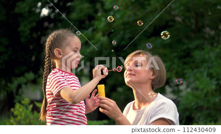 Little girl and mother blow soap bubbles together in green city park. Mother with joyful little girl plays soap bubbles walking in park. Little girl and mother rest blowing soap bubbles in garden 112430409