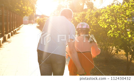 Cute little girl in protective helmet riding on cycle with father at sunlight city park back view closeup. Family man and female kid child driving bike bicycle together weekend childhood sunny outdoor Cute little girl in protective helmet riding on cycle with father at sunlight city park back view closeup. Family man and female kid child driving bike bicycle together weekend childhood sunny outdoor 112430432