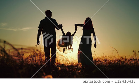 Daughter jumps happily walking across field and tells parents about day at kindergarten. Young daughter boasts of fond memories from kindergarten before walking as family strolls in field at sunset 112430435