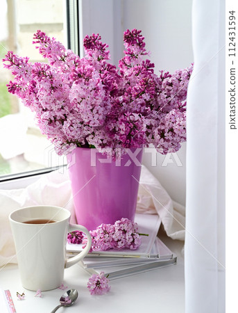 The postcard is beautiful. A mug of tea, an old book and a bouquet of purple lilac. A beautiful still life. Spring time. The concept of Good morning. The postcard is beautiful. A mug of tea, an old book and a bouquet of purple lilac. A beautiful still life. Spring time. The concept of Good morning. 112431594
