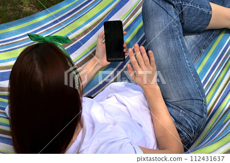 A young happy woman sitting in a park on the green grass in nature with a phone in her hands in casual clothes and chatting by video link, the concept of healthy recreational meditation A young happy woman sitting in a park on the green grass in nature with a phone in her hands in casual clothes and chatting by video link, the concept of healthy recreational meditation 112431637
