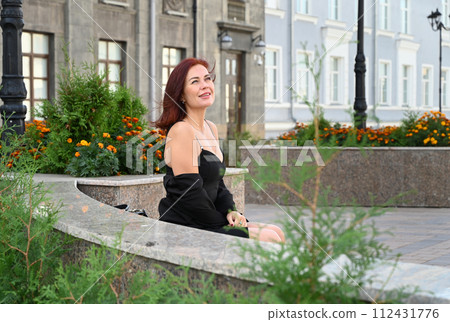 a confident young attractive woman in a black dress and high-heeled shoes walks through the streets of the city, the woman smiles and enjoys life a confident young attractive woman in a black dress and high-heeled shoes walks through the streets of the city, the woman smiles and enjoys life 112431776