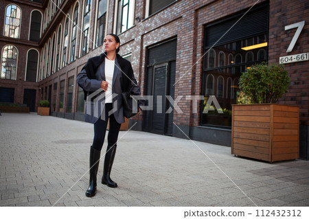 Stylish fashion woman Standing the street in multilayer clothes oversized blazer, black leggings, and knee high rubber boots, large bag. Fashionable outfit, street style Stylish fashion woman Standing the street in multilayer clothes oversized blazer, black leggings, and knee high rubber boots, large bag. Fashionable outfit, street style 112432312