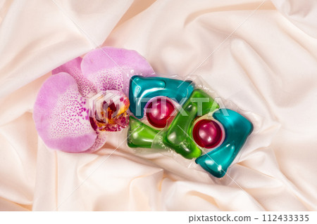 Laundry capsules among orchid flowers on satin fabric. Laundry capsules among orchid flowers on satin fabric. 112433335