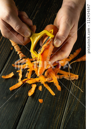 Close-up of a chef peeling raw carrots with a vegetable peeler to prepare a vegetable dish. Delicious carrot breakfast or smoothie. Place for advertising 112434847