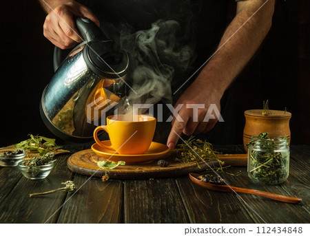 A man brews tea with medicinal herbs on the kitchen table. Traditional medicine concept at home. Adding boiling water to a mug 112434848
