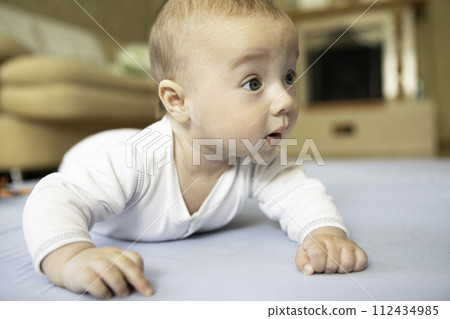 Cute baby lies on his tummy in bright room house, looking with interest various objects around him.  112434985