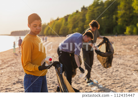 Earth day. Volunteers activists team collects garbage cleaning of beach coastal zone. Woman puts plastic bottle trash in garbage bag on ocean shore. Environmental conservation coastal zone cleaning Earth day. Volunteers activists team collects garbage cleaning of beach coastal zone. Woman puts plastic bottle trash in garbage bag on ocean shore. Environmental conservation coastal zone cleaning 112435447