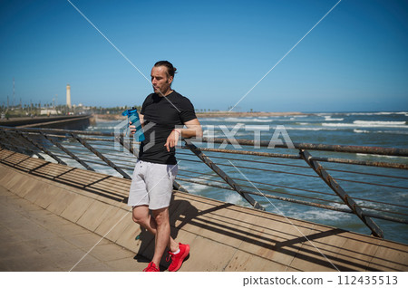 Sportsman with a bottle of water on the beach after morning jog or bodyweight training on the outdoor sportsground 112435513