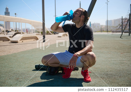 Active athletic young man drinking water from sports bottle while resting in the sportsground after workout outdoors Active athletic young man drinking water from sports bottle while resting in the sportsground after workout outdoors 112435517