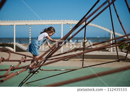 Adorable little girl climbing on a climbing net at the city playground on the embankment. Adorable little girl climbing on a climbing net at the city playground on the embankment. 112435533
