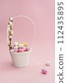 White Easter basket with chocolate multicolored eggs and willow branch on pastel pink background. Easter composition. Card with copy space for text 112435895