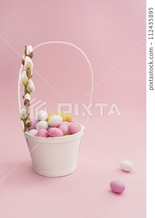 White Easter basket with chocolate multicolored eggs and willow branch on pastel pink background. Easter composition. Card with copy space for text White Easter basket with chocolate multicolored eggs and willow branch on pastel pink background. Easter composition. Card with copy space for text 112435895