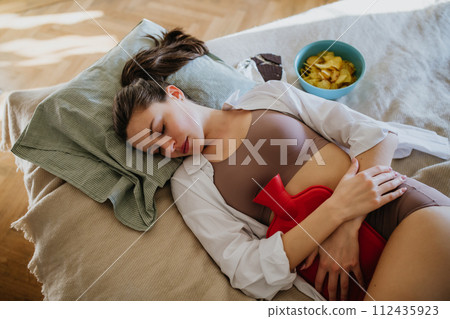 Woman lying on bed with menstrual pain and cramps, having period cravings. Woman warming lower abdomen with a hot water bottle, endometriosis, and conditions causing pain in tummy. Woman lying on bed with menstrual pain and cramps, having period cravings. Woman warming lower abdomen with a hot water bottle, endometriosis, and conditions causing pain in tummy. 112435923