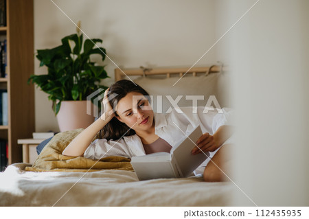 Beautiful woman reading book in bed, cozy, warm and contented feeling. Freedom in story, weekend activity for single woman. 112435935