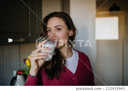 Beautiful woman drinking glass of plant-based milk in the kitchen. 112435941