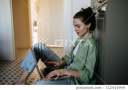 Young student e-learning on laptop at home. Businesswoman working remotely, sitting at kitchen floor, typing. Homeoffice for single woman living alone. 112435944