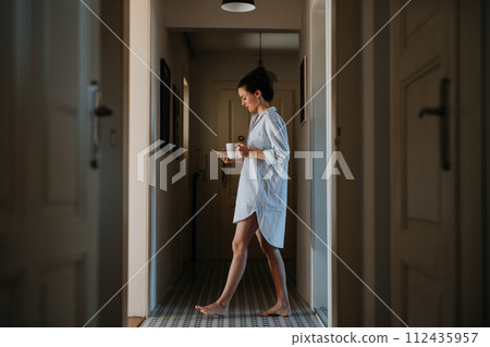 Young woman in the morning, carrying cup of coffee and returning back to bed. Side view of beautiful single woman at home alone over the weekend. Young woman in the morning, carrying cup of coffee and returning back to bed. Side view of beautiful single woman at home alone over the weekend. 112435957