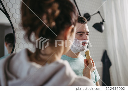 Mother applying shaving foam on son's face. Young man with down syndrome learning how to shave. 112436002