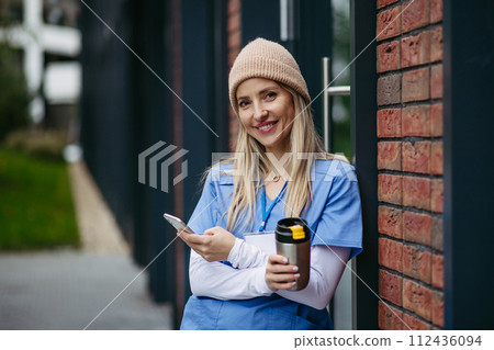 Nurse, doctor in uniform taking break, standing in front hospital building, scrolling on smartphone. Work-life balance of healthcare worker. 112436094