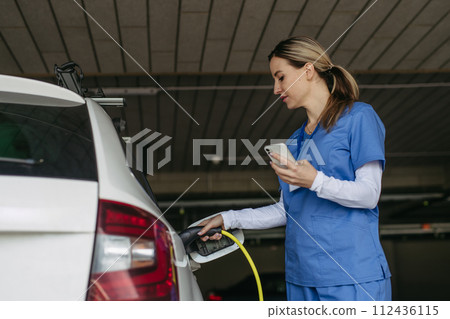 Nurse plugging charger into electric car before going to work. Electric vehicle charging station in front of hospital, modern clinic. Charging at work, workplace. Nurse plugging charger into electric car before going to work. Electric vehicle charging station in front of hospital, modern clinic. Charging at work, workplace. 112436115