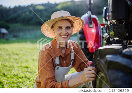 Female farmer fixing a wheel on a tractor. Progressive farmer working on tractor on her own farm. Women in agriculture. 112436269