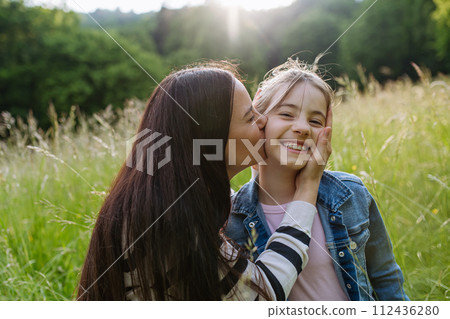 Beautiful mother with daughter, embracing, kissing on cheek, sitting in the grass at meadow. Concept of Mother's Day and maternal love. 112436280