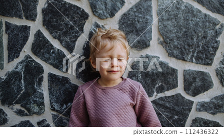 Portrait of little cute girl posing in front of stone wall, eyes closed, not looking at camera. 112436322