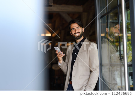 Portrait of mature businessman in suit jacket standing indoors in modern office, holding smartphone. Portrait of mature businessman in suit jacket standing indoors in modern office, holding smartphone. 112436695