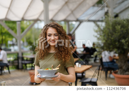 Portrait of a beautiful waitress holding plate with food, bowl with salad. Server standing on restaurant terrace in an apron. Portrait of a beautiful waitress holding plate with food, bowl with salad. Server standing on restaurant terrace in an apron. 112436706