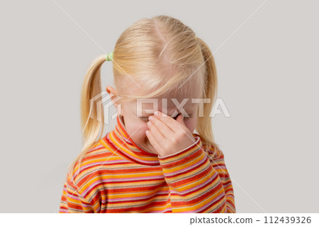 Tired little blonde girl covered her nose with her hand. Isolated on a gray background. Tired little blonde girl covered her nose with her hand. Isolated on a gray background. 112439326