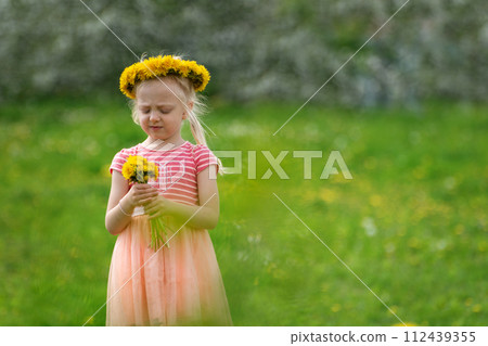 Thoughtful little girl on meadow with flowers. Fair-haired girl wears wreath of dandelions. Summer vacation time. Thoughtful little girl on meadow with flowers. Fair-haired girl wears wreath of dandelions. Summer vacation time. 112439355