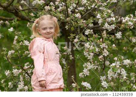 Portrait of cute beautiful blonde girl with two ponytails wears pink jacket stands in spring garden near flowering tree. Portrait of cute beautiful blonde girl with two ponytails wears pink jacket stands in spring garden near flowering tree. 112439359