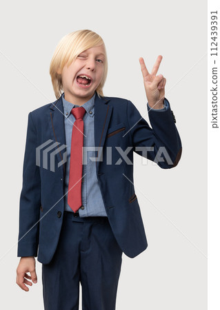 Young squinted Caucasian boy with blond hair, dressed in a blue suit and red tie, showing victory sign with both hands. 112439391