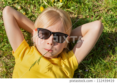 Close up portrait of little blond boy wearing yellow t-shirt and sunglasses lies on grass. Child rests on dandelions filed with his hands behind head. Close up portrait of little blond boy wearing yellow t-shirt and sunglasses lies on grass. Child rests on dandelions filed with his hands behind head. 112439419