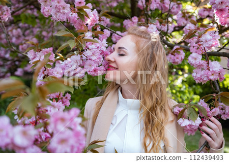 Portrait of happy beautiful woman with wavy hair in blooming park. Attractive smiling girl in white shirt stands near sakura. 112439493