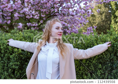 Portrait of attractive woman with curly hair with raised arms in blooming spring park. Girl outdoor, blooming sakura tree background. 112439508