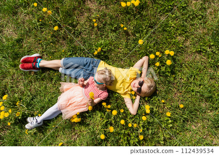 Children boy and younger sister are lying on green field with dandelions. Top view. Siblings rest in the grass. Children boy and younger sister are lying on green field with dandelions. Top view. Siblings rest in the grass. 112439534