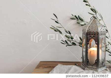 Ramadan Kareem, Eid ul Fitr holiday still life. Ornamental silver Moroccan lantern with olive tree branches on wooden table, bench. Burning candle. Blurred white wall background. Iftar decor. Ramadan Kareem, Eid ul Fitr holiday still life. Ornamental silver Moroccan lantern with olive tree branches on wooden table, bench. Burning candle. Blurred white wall background. Iftar decor. 112439637