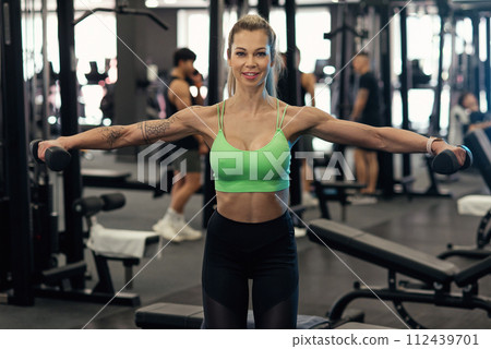 Muscular fitness woman athlete doing heavy weight exercise for shoulders with dumbbells in the gym 112439701