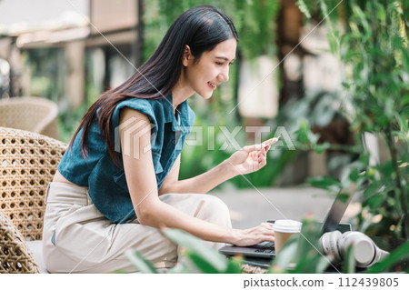 A smiling woman sits comfortably outdoors with a laptop and headphones, holding a credit card, ready to complete an online transaction in a tranquil garden environment. A smiling woman sits comfortably outdoors with a laptop and headphones, holding a credit card, ready to complete an online transaction in a tranquil garden environment. 112439805