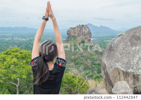 Cropped shot view of young woman doing Yoga on top of Pidurangala rock and looking to Sigiriya rock an iconic tourist destination and one of UNESCO world heritage site in Sri Lanka. 112442847