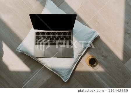 Photo of laptop showing black empty screen putting on white pillow, on wooden floor in cozy room with black coffee cup. Photo of laptop showing black empty screen putting on white pillow, on wooden floor in cozy room with black coffee cup. 112442889