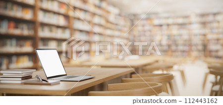 A white-screen laptop computer mockup and books on a wooden table in a spacious university library. 112443162