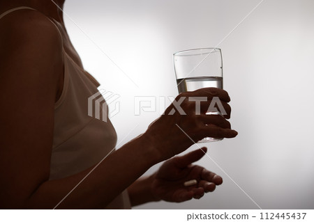 Silhouette of a woman holding a glass of water and drinking medicine by the window 112445437